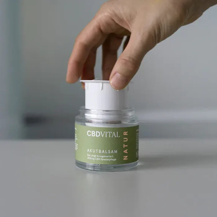 Eine Hand nimmt den weißen Refill-Einsatz aus dem CBD Vital Akutbalsam Glastiegel