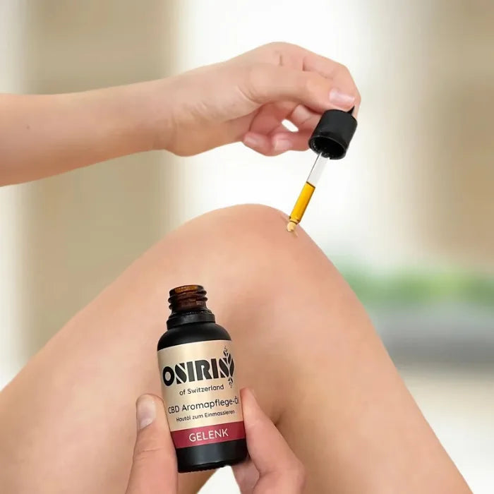 Osiris Aromapflege Öl Gelenk und Muskel, ein Tropfen Hautöl fällt aus der Pipette auf die Haut am Knie, Flasche in der anderen Hand