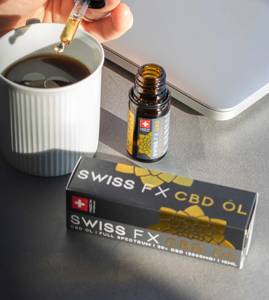 Hand fügt Swiss FX CBD Öl mit einem Tropfer zu einer Tasse Kaffee hinzu, neben der geöffneten Produktverpackung und einer Flasche auf einem Tisch mit Laptop im Hintergrund