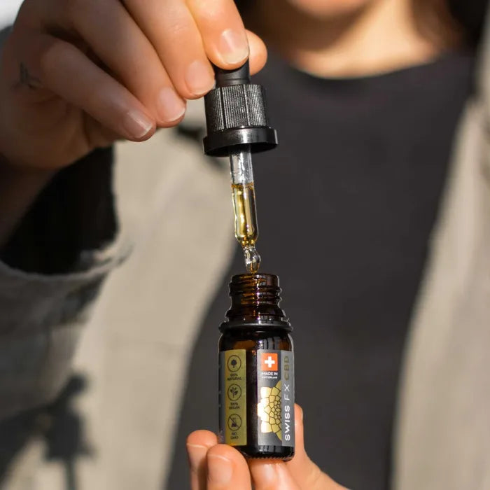 Hand hält die Pipette über die geöffnete Swiss FX CBD Pure Flasche aus Braunglas, ein Tropfen CBD Öl fällt in die Flasche